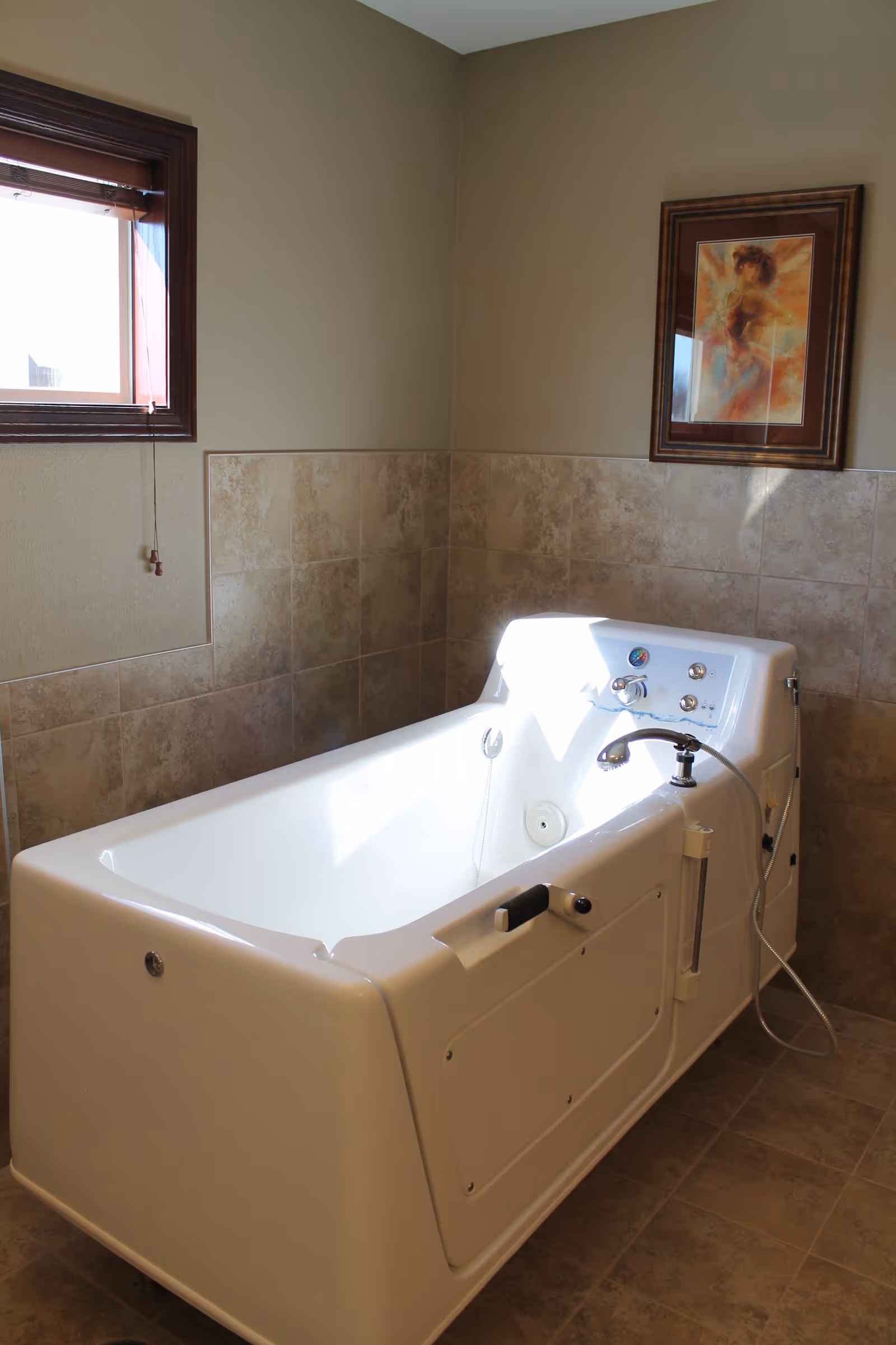 A white walk-in bathtub with control knobs and a handheld shower head in a bathroom with beige tiled walls and floor. There is a small window with wooden trim and a framed artwork on the wall above the tub.