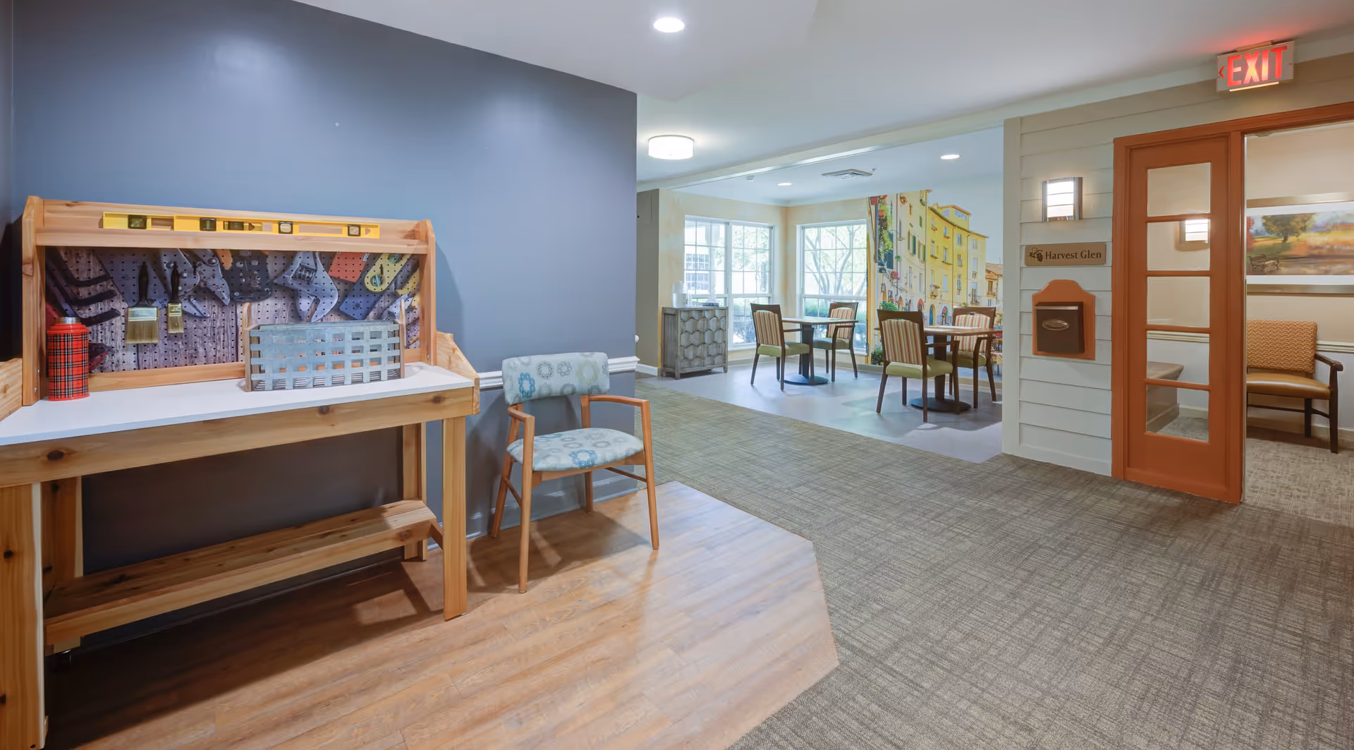 Interior view of a senior living facility hallway with a wooden activity table on the left, a patterned chair next to it, and a dining area with tables and chairs in the background. The walls are painted in neutral tones, and there is an exit sign above a door on the right side.