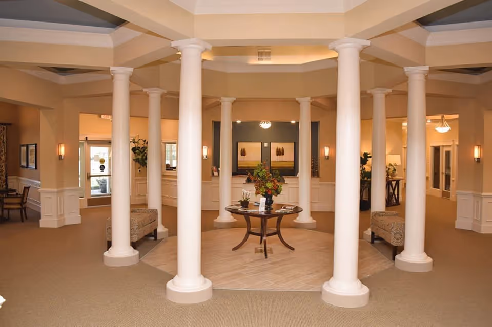 Interior view of a senior living facility lobby area featuring a circular arrangement of white columns surrounding a round wooden table with a floral centerpiece. The space is warmly lit with beige walls, carpeted floors, and wall sconces. There are seating areas and doorways visible in the background.