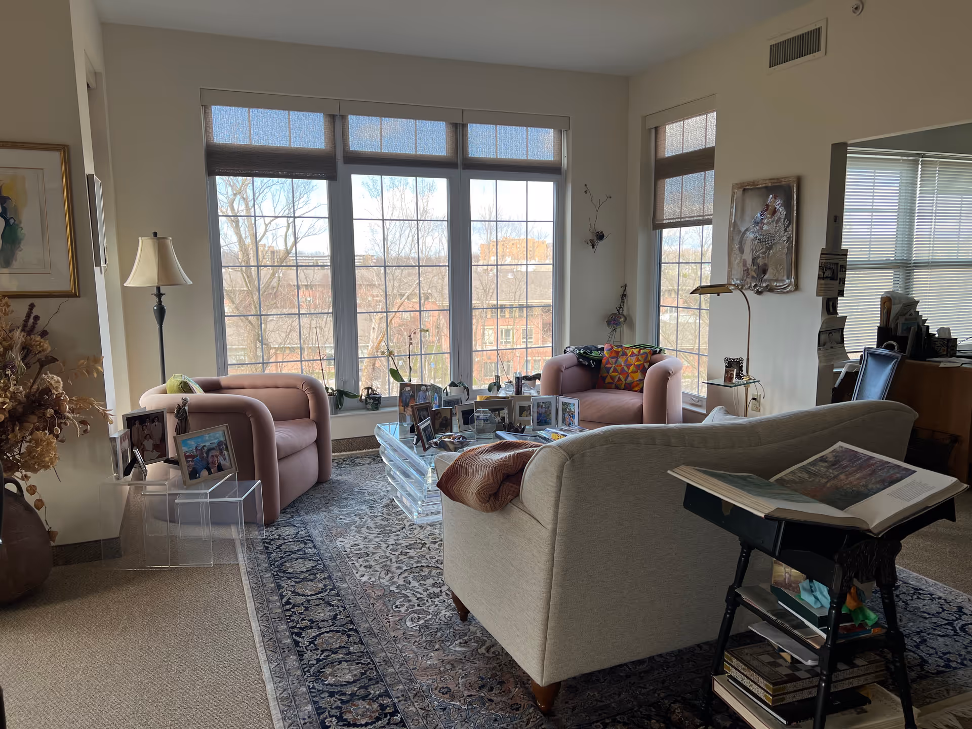A cozy living room with large windows letting in natural light. The room features two pink armchairs, a beige sofa, a glass coffee table with framed photos, a floor lamp, and a side table with a large open book. There is a patterned rug on the floor and artwork on the walls.
