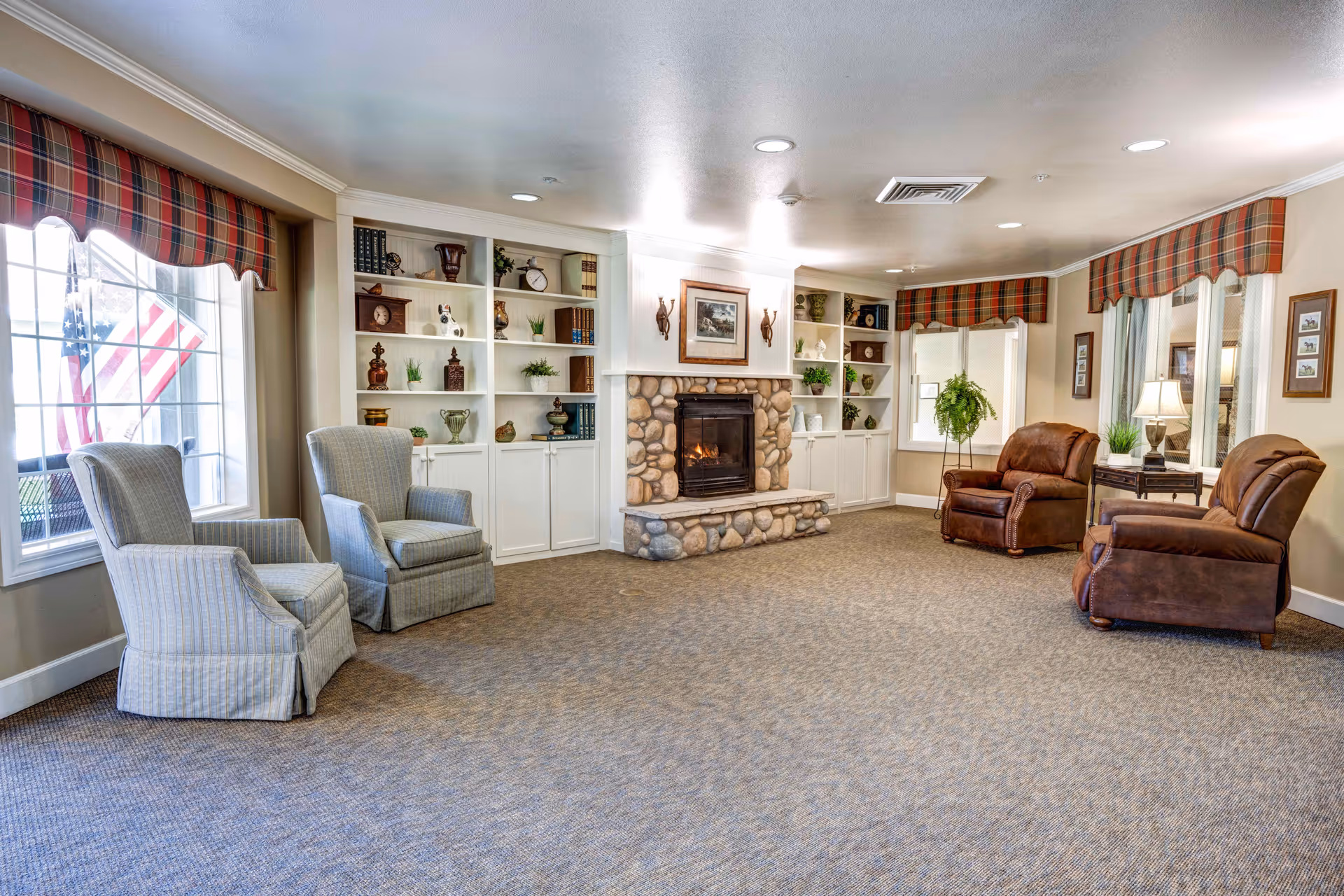 Bright communal living room with a stone fireplace, built-in shelves, upholstered armchairs and leather recliners, and large windows with plaid valances.