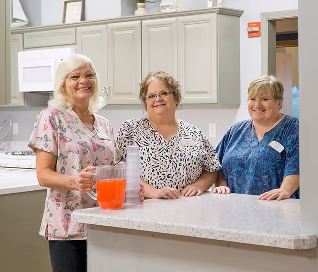 Three smiling caregivers standing behind a kitchen counter in a memory care facility with a pitcher of red drink and stacked plastic cups.