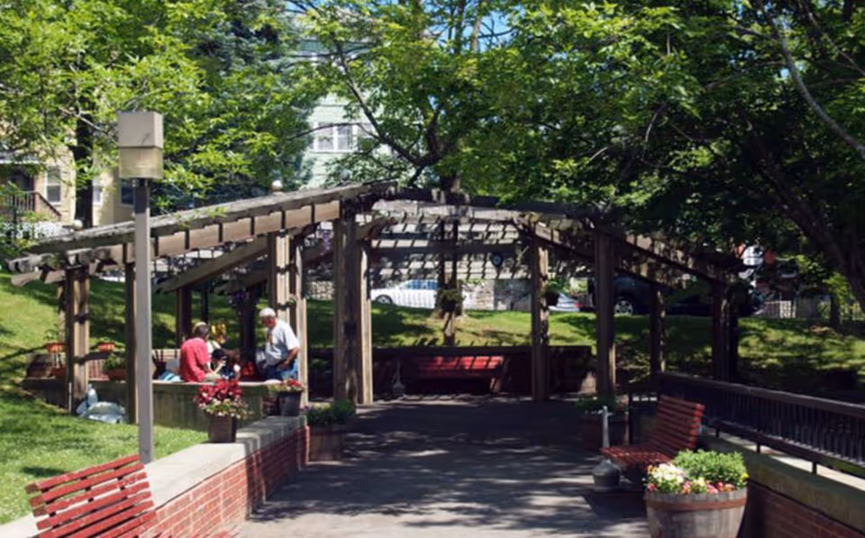 Outdoor garden area with a wooden pergola structure providing partial shade. Several people are seated and standing under the pergola, surrounded by green trees and grass. There are red benches and flower pots along the paved pathway.