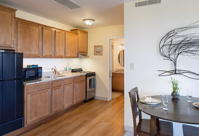 Interior view of a kitchen and dining area in a senior living facility. The kitchen features wooden cabinets, a black refrigerator, microwave, sink, and a stove with an oven. Adjacent to the kitchen is a small dining table set with plates, glasses, and napkins. A decorative metal wall art piece is mounted on the wall above the dining table. In the background, a doorway leads to a bathroom with a vanity and mirror.