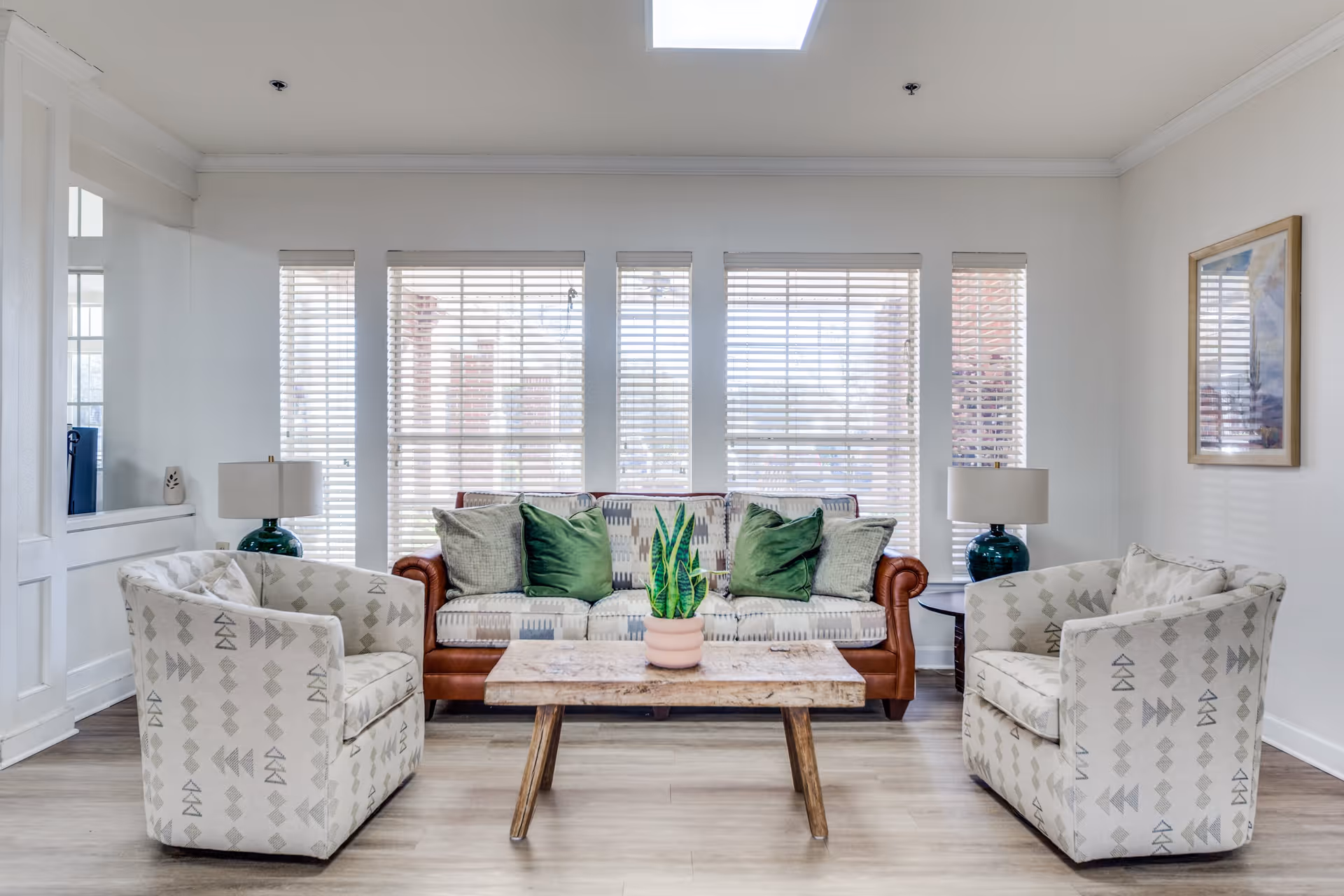 A bright and cozy living room with a patterned sofa adorned with green and beige cushions, flanked by two matching armchairs. A wooden coffee table with a potted plant sits in the center. Large windows with white blinds allow natural light to fill the room. Two table lamps with green bases are placed on side tables beside the sofa. A framed picture hangs on the wall to the right.