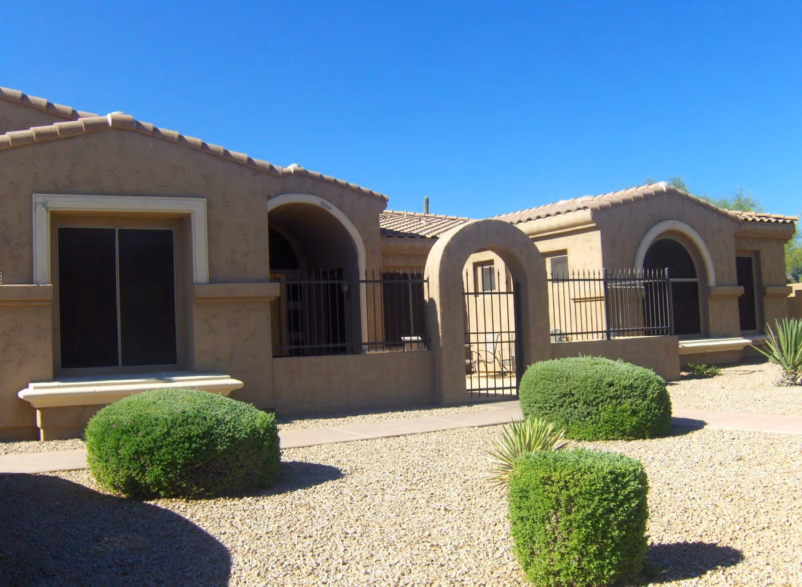 Exterior view of a single-story building with beige stucco walls and a tiled roof under a clear blue sky. The building features arched doorways and windows with black metal fencing enclosing a small patio area. The foreground has a gravel landscape with neatly trimmed green bushes and a paved walkway leading to the gated entrance.