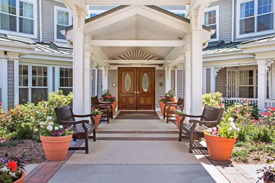 Entrance to a senior living facility with a covered walkway, wooden double doors, benches on either side, and large flower pots with colorful flowers. The building exterior is light gray with white trim and multiple windows.
