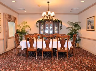 A formal dining room with a long wooden table covered with a white tablecloth and surrounded by eight wooden chairs. A chandelier hangs above the table. There is a wooden china cabinet with glass doors and decorative greenery on top against the back wall. Two large potted plants are placed on either side of the cabinet. The room has patterned carpet, beige walls with a chair rail, and windows with draped curtains on the left side.