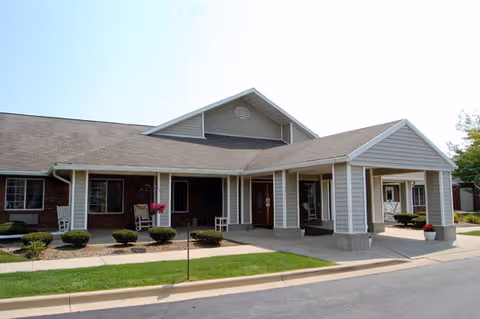 Exterior view of a single-story senior living facility building with a covered entrance, white rocking chairs on the porch, small bushes, and a well-maintained lawn under a clear sky.