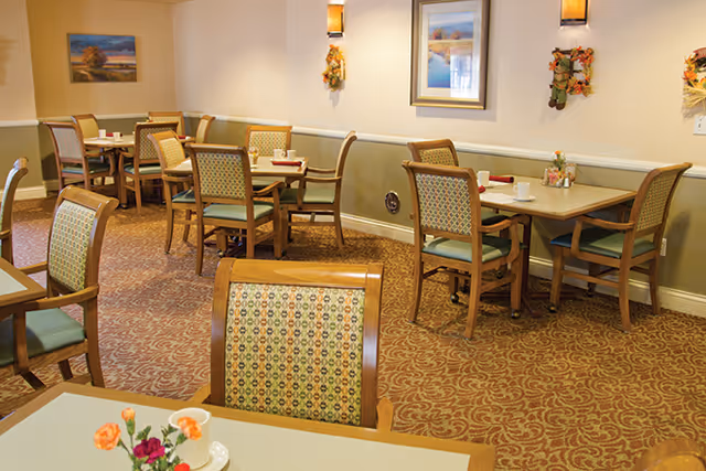 Dining room with multiple small tables and patterned wooden chairs, floral centerpieces, and wall artwork.