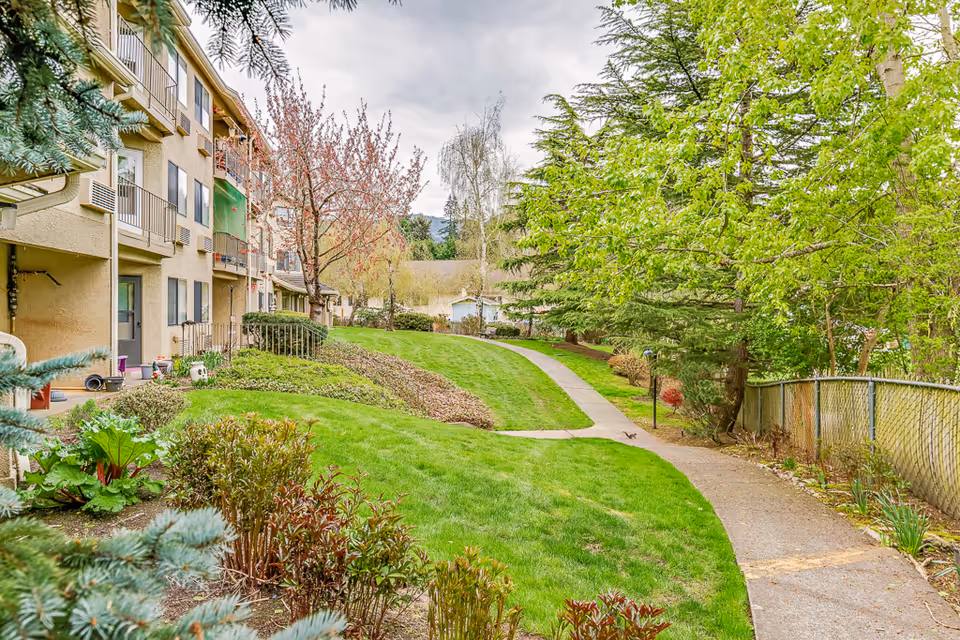 Outdoor view of a senior living facility showing a well-maintained grassy area with a paved walking path. On the left side, there is a multi-story building with balconies and windows. The area is surrounded by various trees and shrubs, creating a peaceful and green environment.