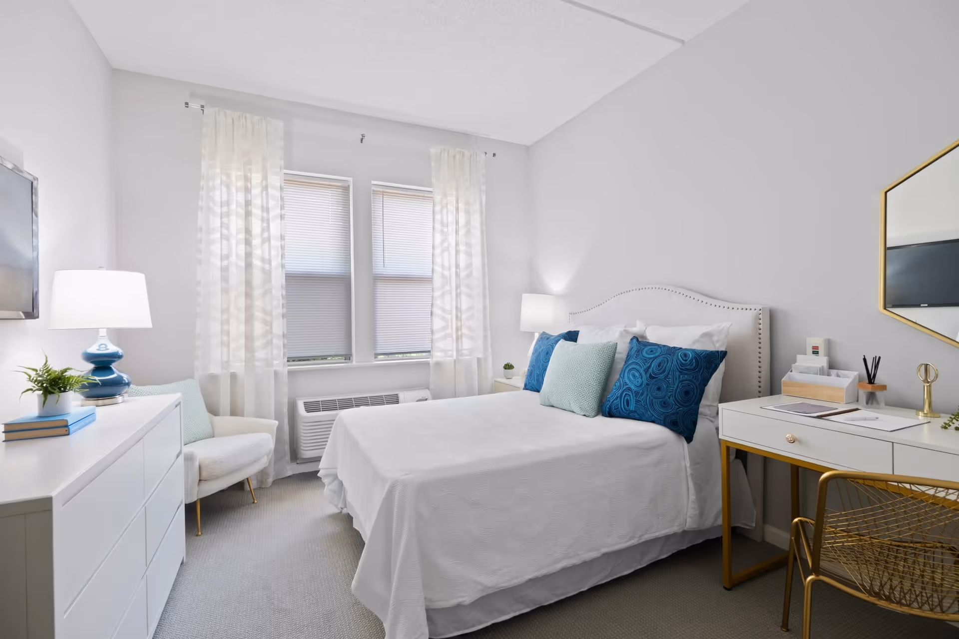 Bright modern bedroom with a white bed topped with blue accent pillows, a desk, dresser, armchair, and two windows with sheer curtains.
