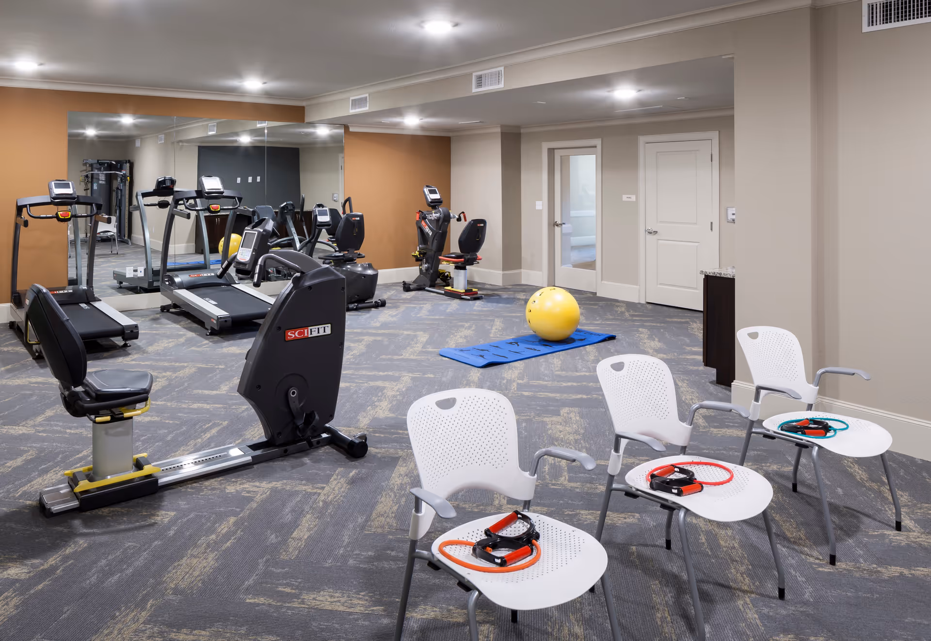 Interior fitness room with treadmills, recumbent bikes, chairs, resistance bands, a yoga mat and a large yellow exercise ball.