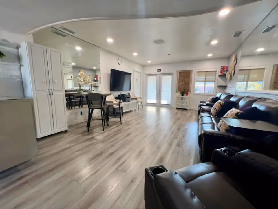 A spacious and modern living room with light wood flooring, a row of dark leather recliners with patterned pillows on the right, a wall-mounted flat screen TV, a small table with two chairs, a large mirror on the left wall, and double glass doors at the far end letting in natural light.