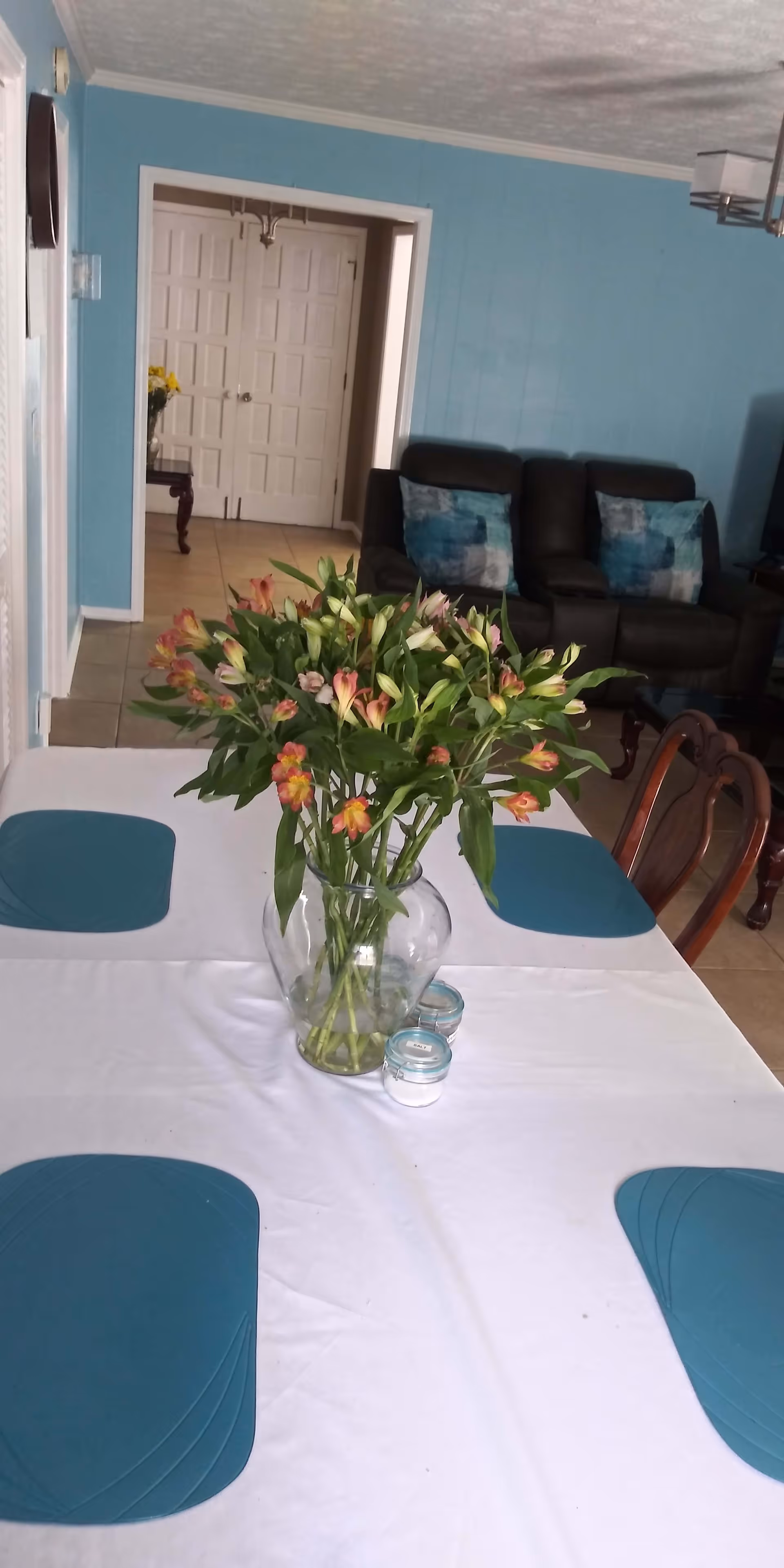 A dining table with a white tablecloth and four teal placemats, centered with a clear glass vase filled with pink and yellow flowers. In the background, there is a living room area with a dark brown recliner sofa with blue patterned pillows, light blue walls, and a doorway leading to another room with double white doors.