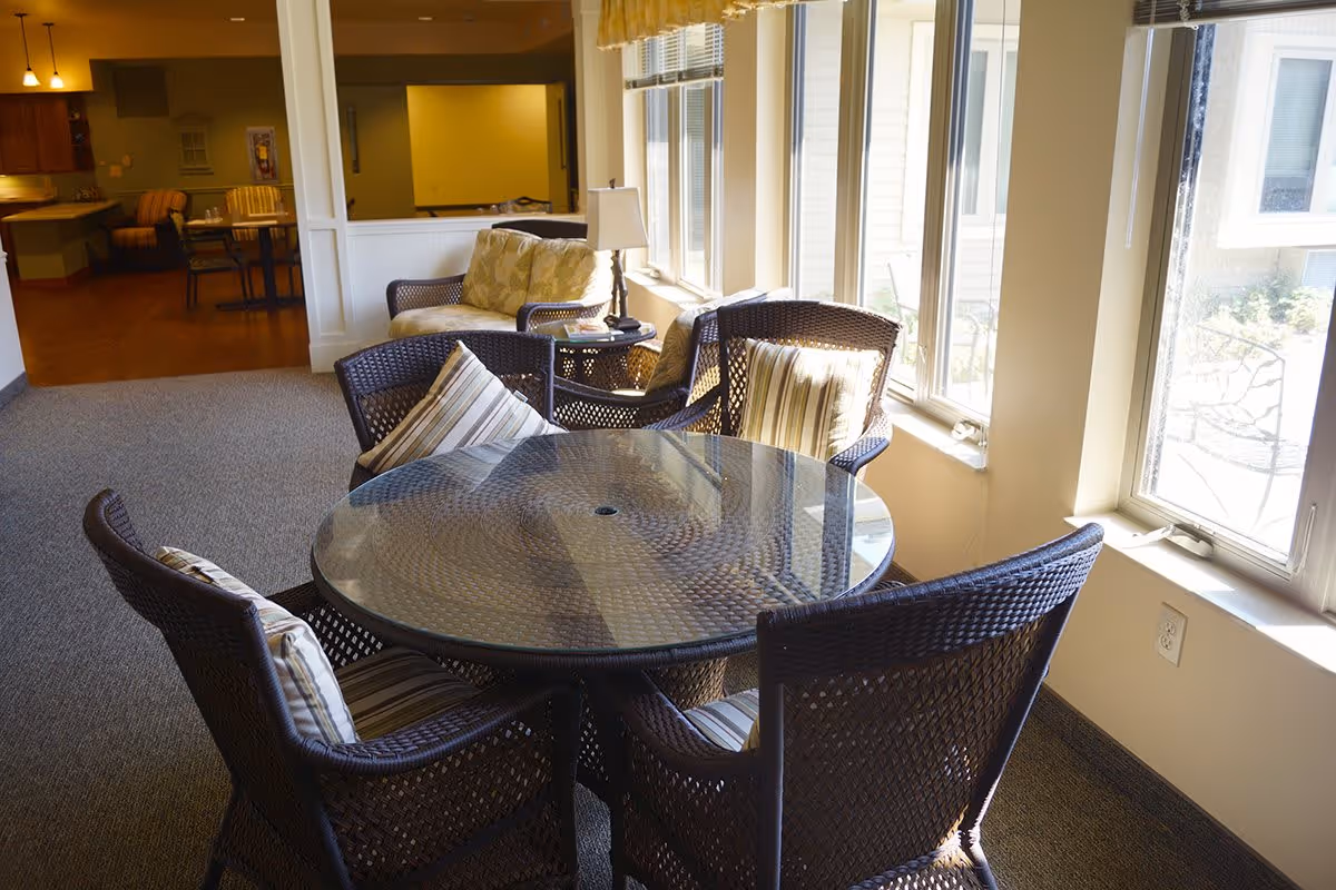 A bright indoor seating area with a round glass-top table surrounded by four wicker chairs with striped cushions. Behind the table, there is a cushioned loveseat and armchair with a small side table and lamp. Large windows on the right side let in natural light, illuminating the carpeted floor and adjacent dining area in the background.
