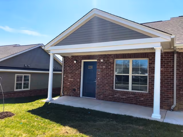 Exterior view of a brick apartment unit with a covered porch supported by two white columns. The unit has a dark blue door with the number 339 and a window to the right of the door. There is a grassy area in front of the porch and another building visible in the background under a clear blue sky.