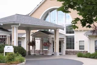 Entrance of a senior living facility named The Pointe at Cascades of Grants Pass, featuring a covered drop-off area with hanging flower baskets, large windows, and beige siding with stone accents.