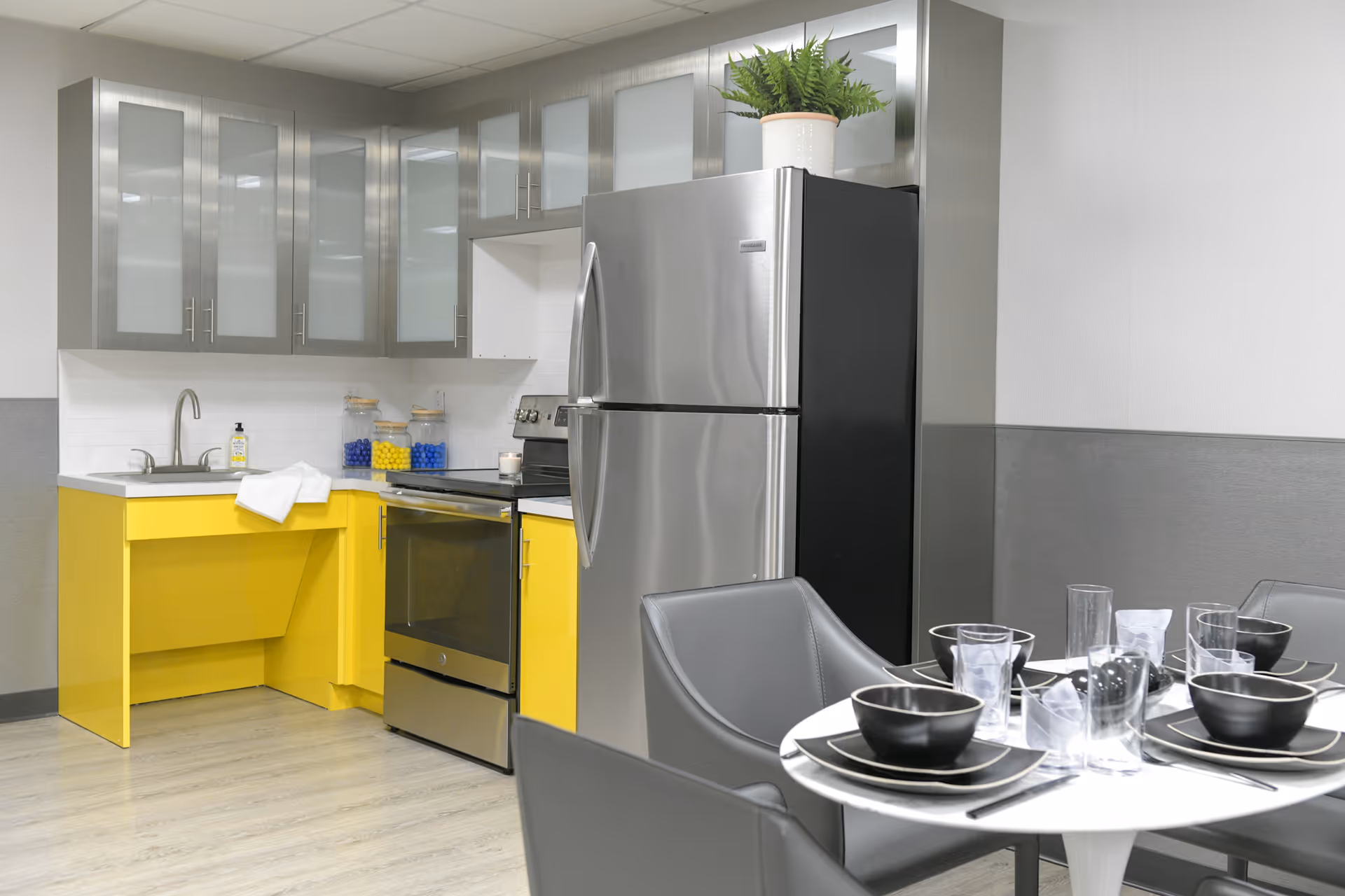 Modern kitchen area with bright yellow lower cabinets and gray upper cabinets with frosted glass doors. Stainless steel refrigerator and oven are present. A small round dining table is set with black bowls, plates, glasses, and silverware. A potted plant is on top of the refrigerator.