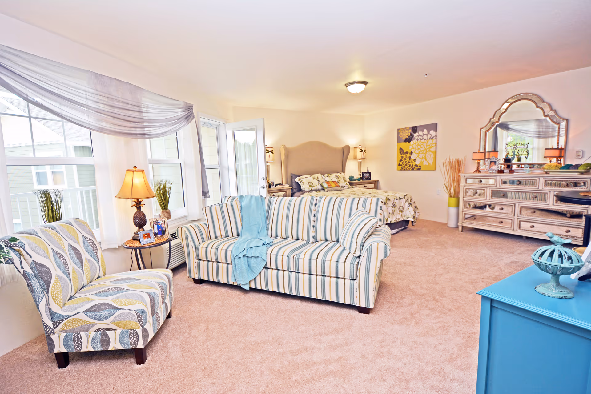 A bright and spacious bedroom with a beige carpet, featuring a striped sofa with a blue throw, a patterned armchair, a bed with a beige headboard and floral bedding, a large dresser with a decorative mirror, and various lamps and decorative items. Large windows with sheer curtains allow natural light to fill the room.