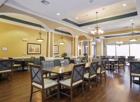 Spacious assisted living dining room with multiple wooden tables and chairs, chandeliers, and large windows.