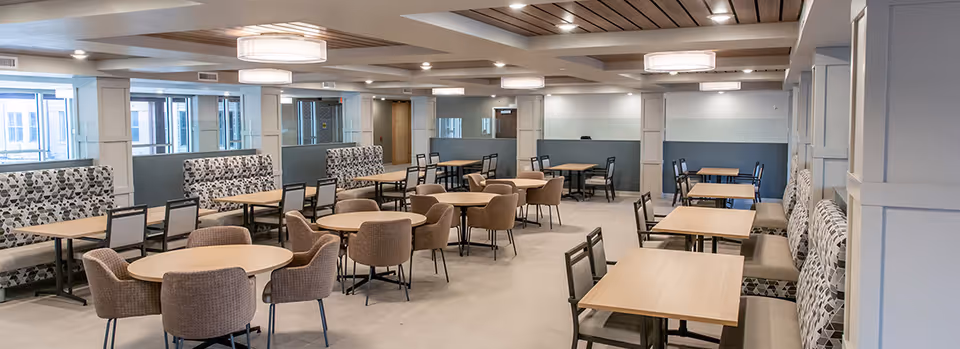 A spacious dining room with multiple tables and chairs arranged neatly. The room features cushioned bench seating along the walls with patterned upholstery, round and square wooden tables, and comfortable padded chairs. Large windows allow natural light to brighten the space, and modern ceiling lights provide additional illumination.