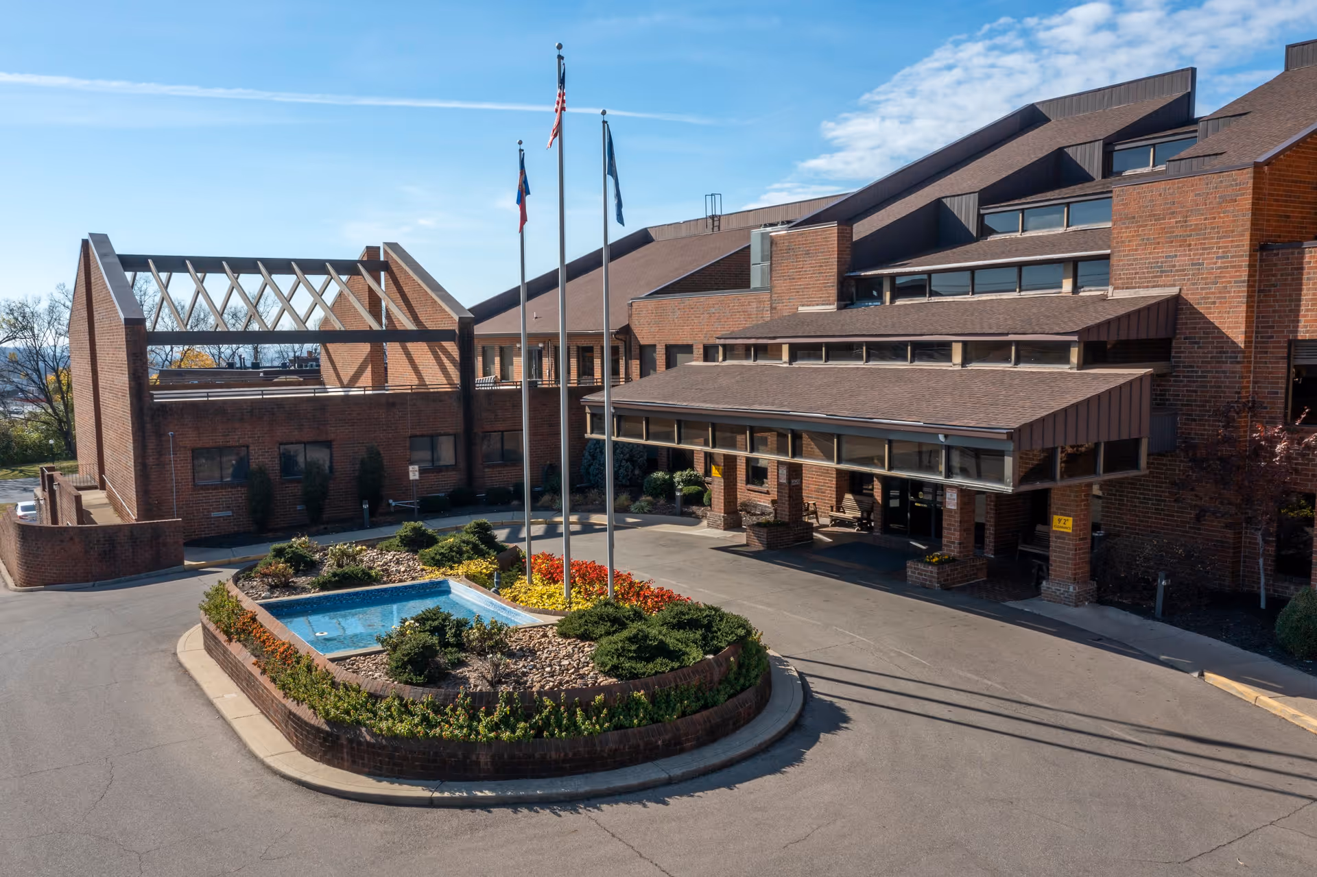 Exterior view of Cottingham Retirement Community building with a driveway, landscaped roundabout featuring a small fountain, shrubs, and flowers, and three flagpoles with flags against a partly cloudy sky.