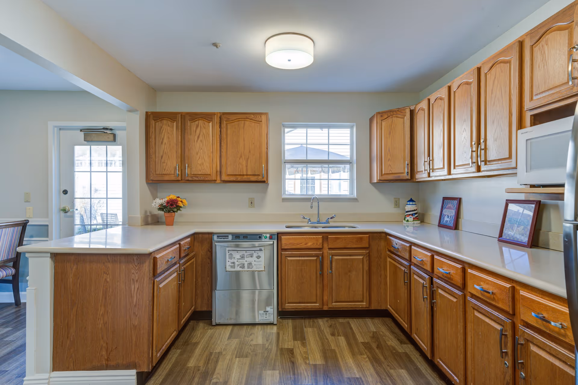 A bright kitchen with wooden cabinets and drawers, a stainless steel dishwasher, a white countertop, and a window above the sink. There is a ceiling light fixture, a microwave, and some decorative items including a small flower pot and framed pictures on the countertop. The kitchen floor is wood, and there is a door leading to another room with a dining table and chairs visible.