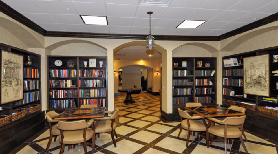 Interior view of a library or reading room with built-in bookshelves filled with books, two wooden tables with four beige cushioned chairs each, decorative framed artwork on the walls, and a tiled floor with a diamond pattern. The room has arched doorways leading to a hallway with warm lighting.