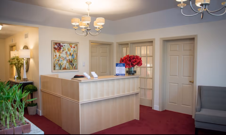 Reception area with a wooden front desk, a vase of red flowers on the desk, a colorful floral painting on the wall, beige doors including one with glass panels, a gray upholstered chair, and indoor plants. The room has a red carpet and ceiling light fixtures.