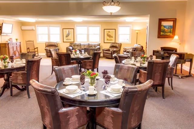 A bright and spacious dining room with multiple round tables set with white dishes, cups, and folded napkins. Each table has a small flower arrangement. The room features comfortable upholstered chairs, a seating area with leather armchairs and a sofa near windows, framed bird artwork on the walls, and a piano in the corner with a lamp beside it.