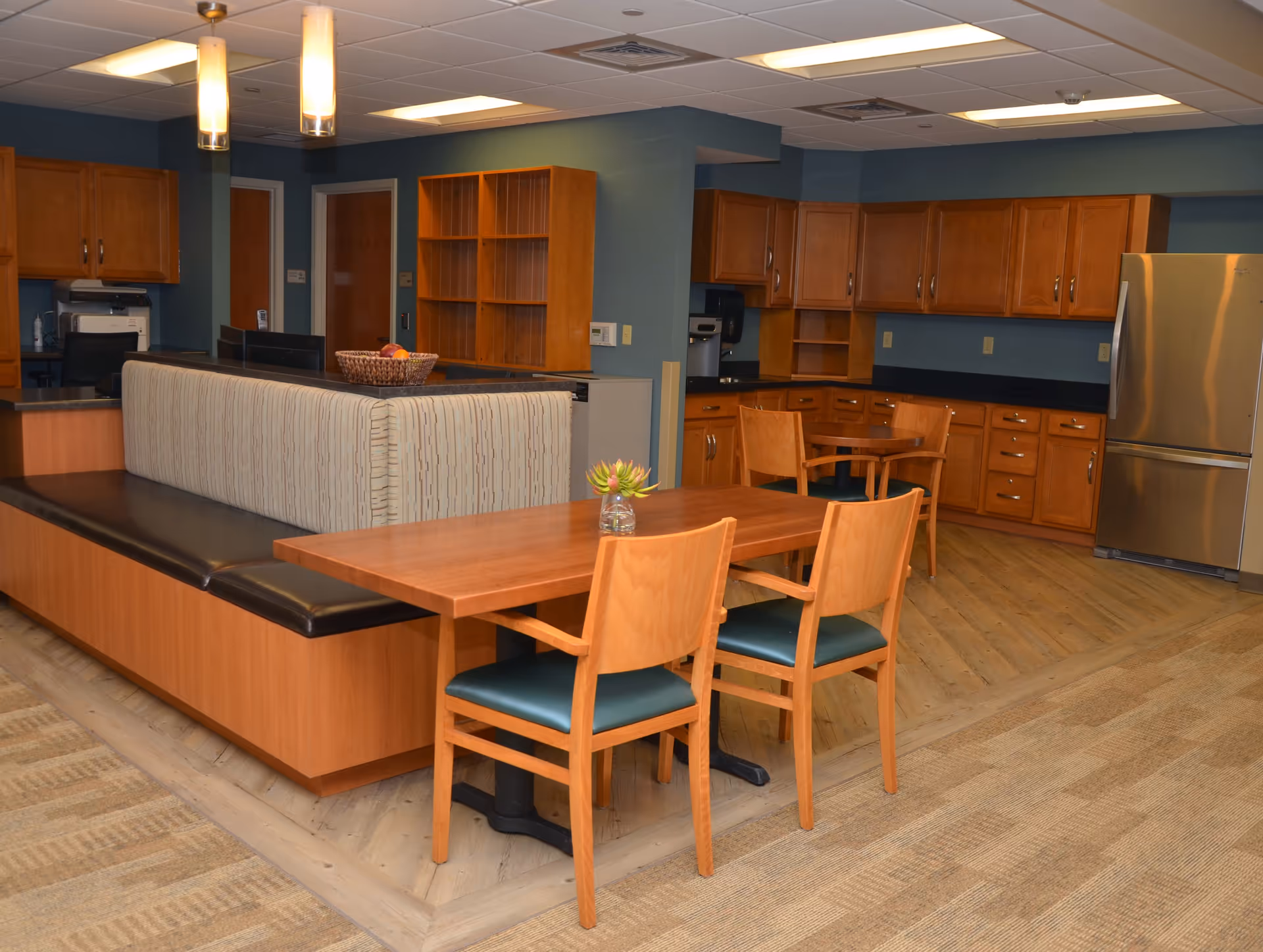 Interior view of a communal dining and kitchen area with wooden tables and chairs, a cushioned bench, wooden cabinets, a stainless steel refrigerator, and overhead lighting fixtures.