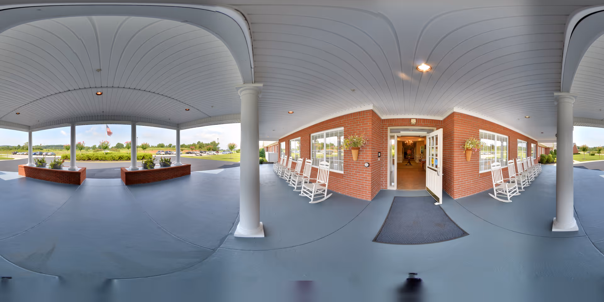 Covered entrance area of The Terrace at Priceville with white columns and ceiling, brick walls, and a row of white rocking chairs along the wall. The entrance door is open, leading inside the building. Outside, there is a parking lot and landscaped greenery with an American flag visible in the distance.