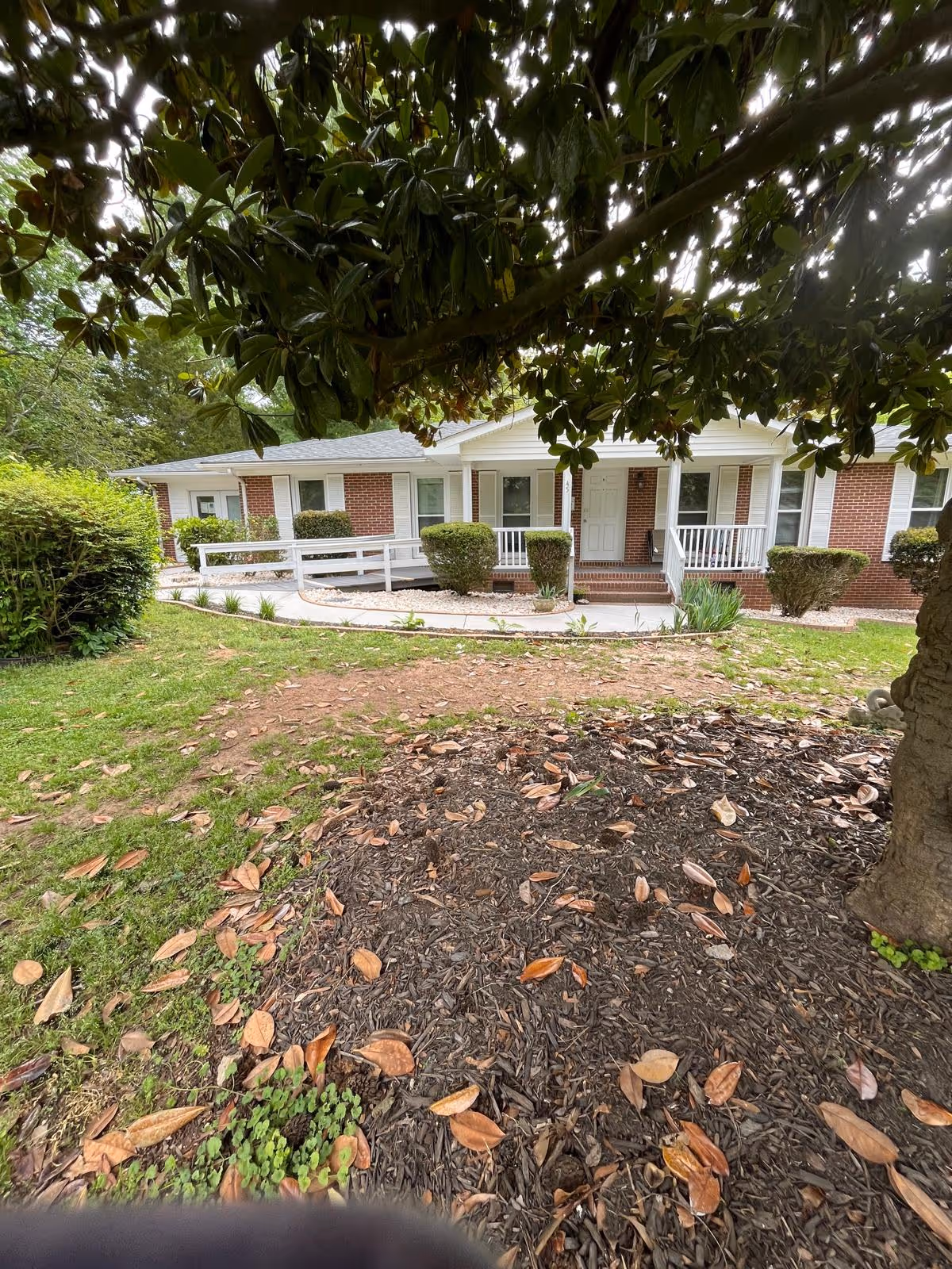 Front exterior view of a single-story brick building with white trim and a covered porch. The porch has white railings and steps leading up to a white door. There are neatly trimmed bushes along the front of the building and a large tree with broad leaves partially shading the view. The foreground shows a mulched area with fallen leaves and some grass.