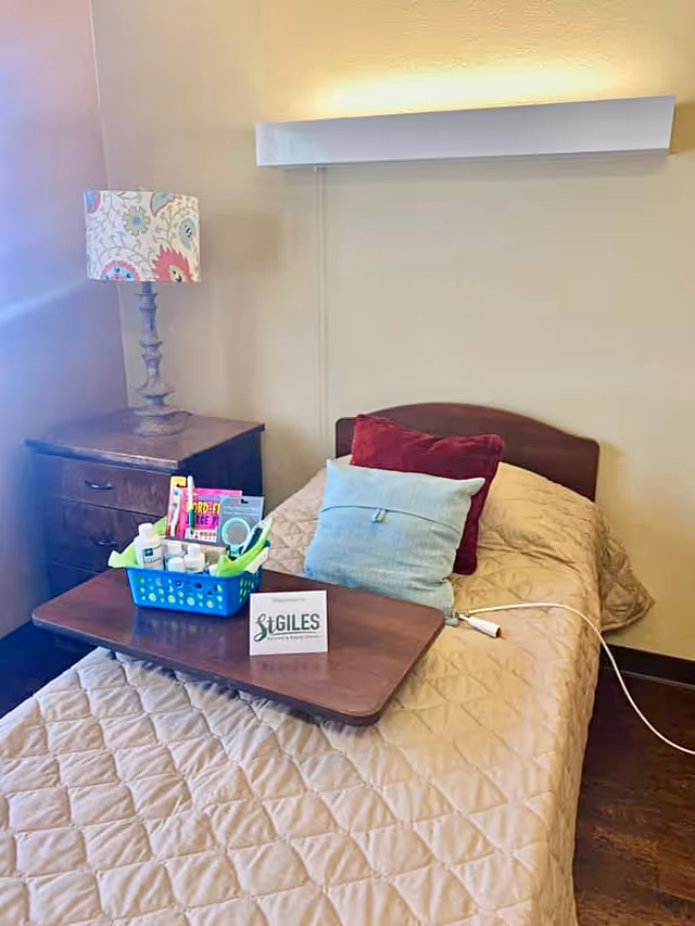 A single bed with a beige quilted bedspread, two pillows (one red and one light blue), and a wooden overbed table holding a blue basket with toiletries and a welcome card from St. Giles Nursing and Rehabilitation Center. Next to the bed is a wooden nightstand with a decorative lamp featuring a floral patterned lampshade. The room has beige walls and a wooden floor.