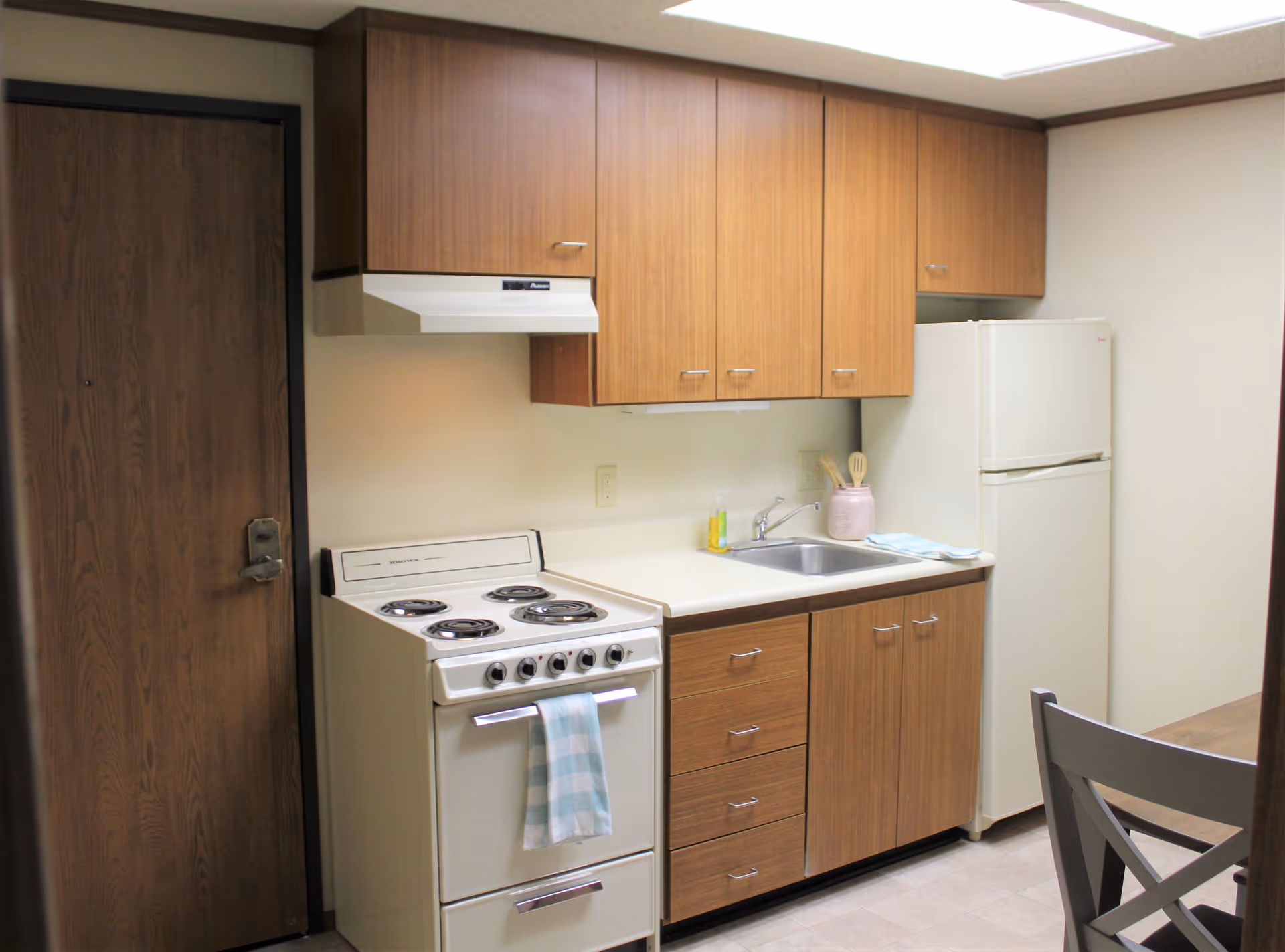 A small kitchen area with wooden cabinets, a white electric stove with four burners and a blue and white checkered towel hanging on the oven handle, a stainless steel sink with a soap dispenser and utensils in a pink container, a white refrigerator, and part of a wooden dining table with a gray chair.