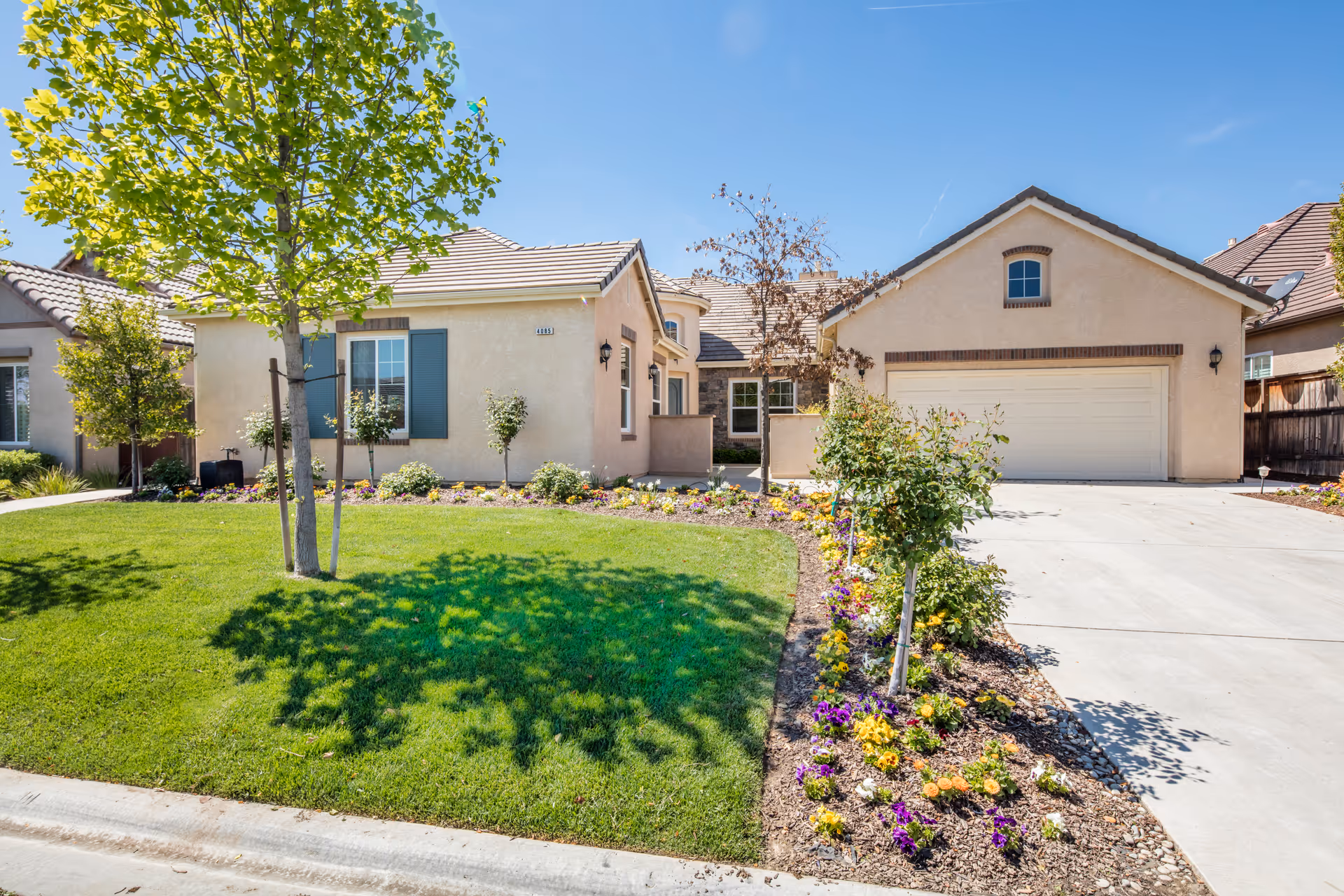 Single-story residential building with an attached garage, green lawn, trees, and flower beds under a blue sky.