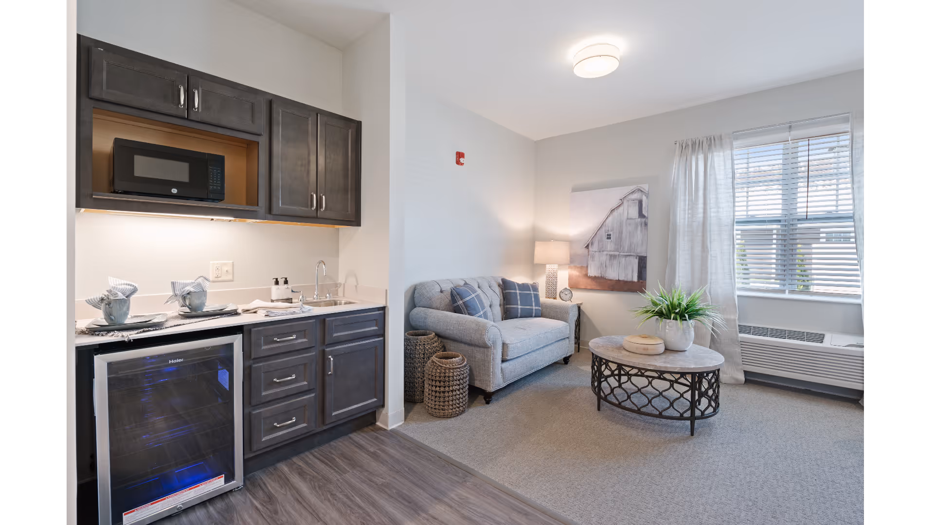 A bright senior living suite with a kitchenette on the left and a loveseat, coffee table, lamp, and window on the right.