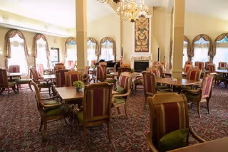 A spacious dining room with multiple wooden tables and upholstered chairs arranged neatly. The room features large windows with decorative curtains, a chandelier hanging from the ceiling, and a fireplace with an ornate tapestry above it. The carpet has a detailed pattern, and the overall ambiance is warm and inviting.