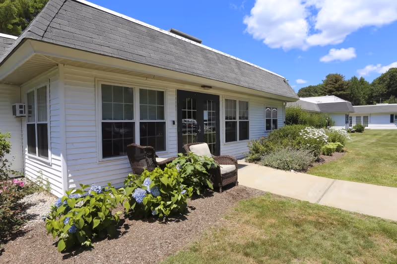 Front exterior of a single-story nursing facility with patio chairs, hydrangea bushes, and a concrete walkway.