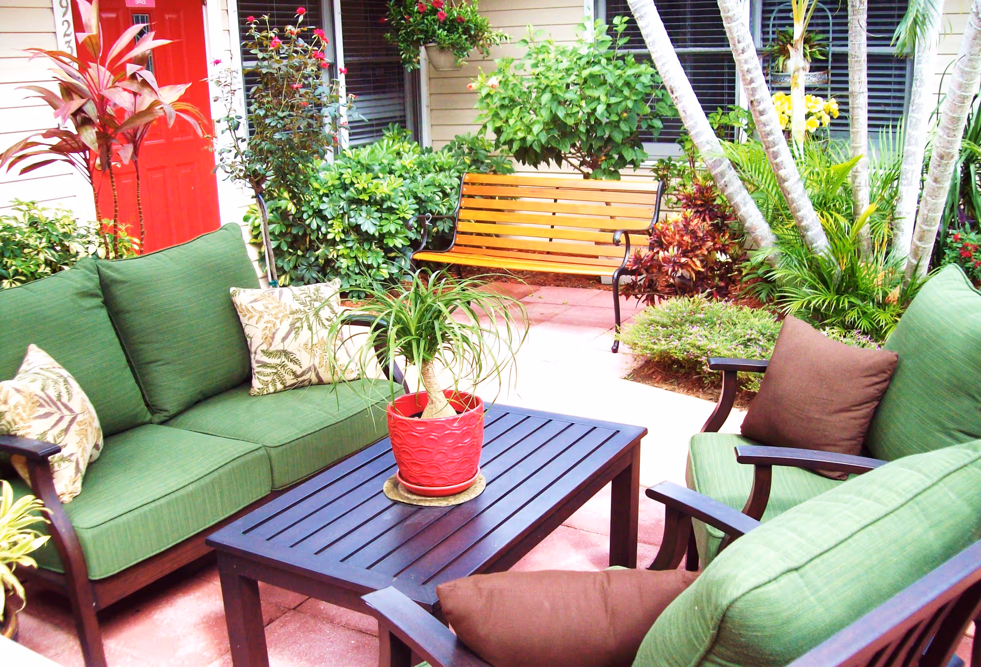 Outdoor patio area with green cushioned chairs and a sofa surrounding a black slatted table with a red potted plant on top. There is a wooden bench in the background, surrounded by various green plants and shrubs, with a red door and windows of a building visible behind.