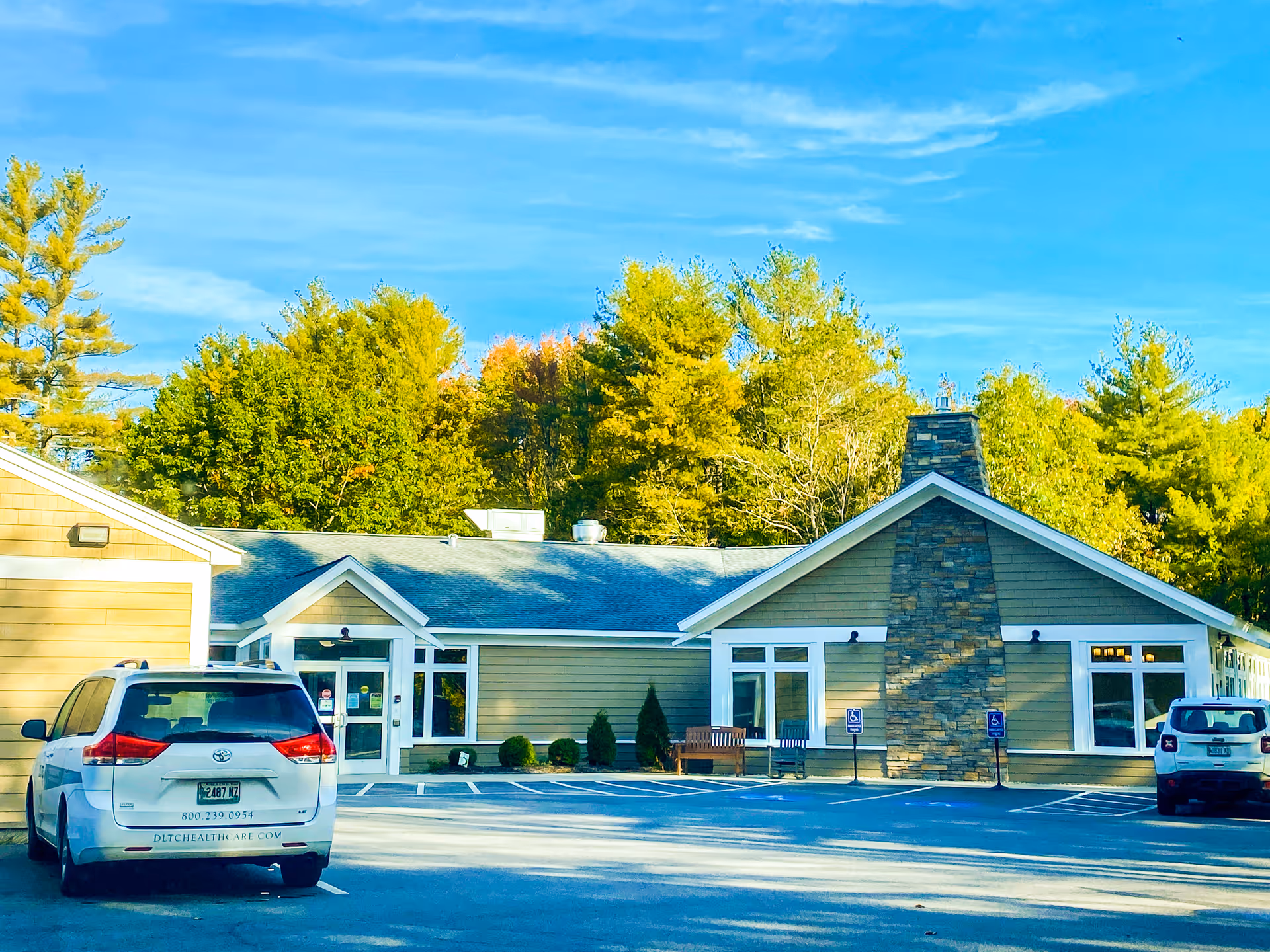 Exterior view of The Lodges Care Center building with a stone chimney, beige siding, and a blue roof. There are two cars parked in front, including a white minivan with healthcare contact information on the back. The building is surrounded by green trees under a clear blue sky.