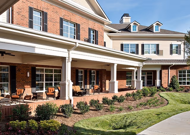 Exterior view of a brick and siding building with a covered porch featuring several cushioned chairs and small tables. The porch is supported by white columns and overlooks a landscaped garden with shrubs and a curved concrete walkway under a clear blue sky.