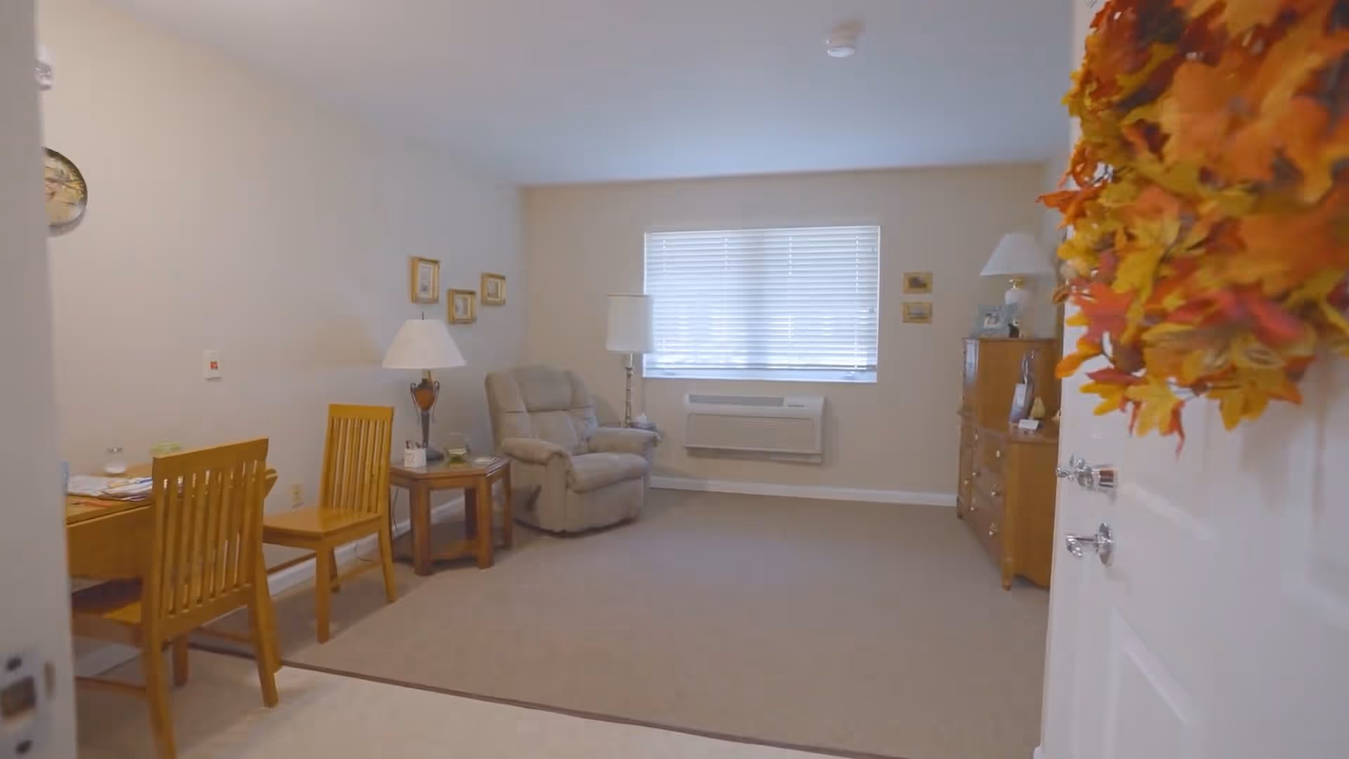 View into a cozy living room area with beige walls and carpet. There is a beige recliner chair next to a window with blinds, a wooden side table with a lamp, and a wooden dresser with a lamp and framed photos. Two wooden chairs and a small table are visible on the left side. The door on the right has a fall-themed wreath hanging on it.