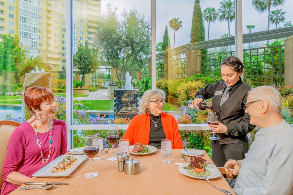 Three older adults seated at a dining table with meals and wine while a server pours wine, with a landscaped garden visible through large windows behind them.
