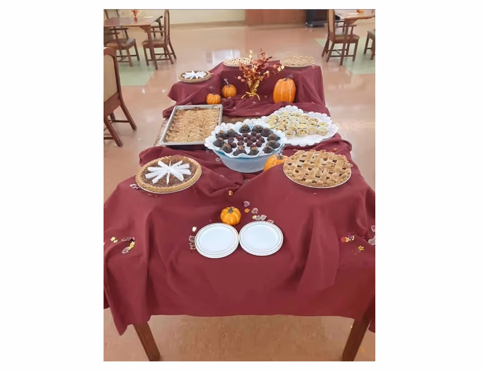 A buffet table in a dining area covered with a burgundy tablecloth displaying pies, pastries, small pumpkins and plates.