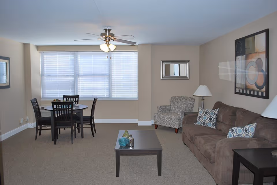 A living room and dining area in a senior living facility with a brown sofa adorned with patterned pillows, a patterned armchair, a coffee table with decorative items, a dining table with four chairs, a ceiling fan with lights, and large windows with blinds.
