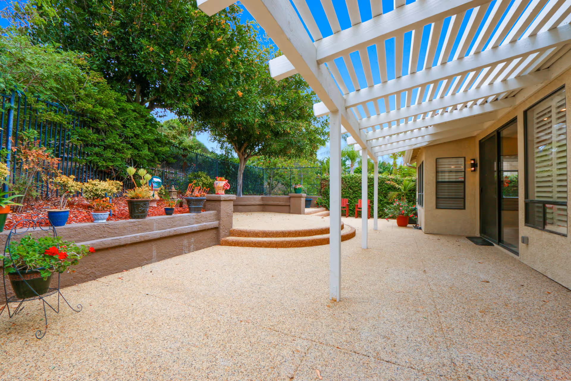 Outdoor patio area with a white pergola overhead, beige textured flooring, and a raised garden bed with various potted plants and flowers. There are trees and shrubs along a black metal fence in the background, and a sliding glass door leading into the building.