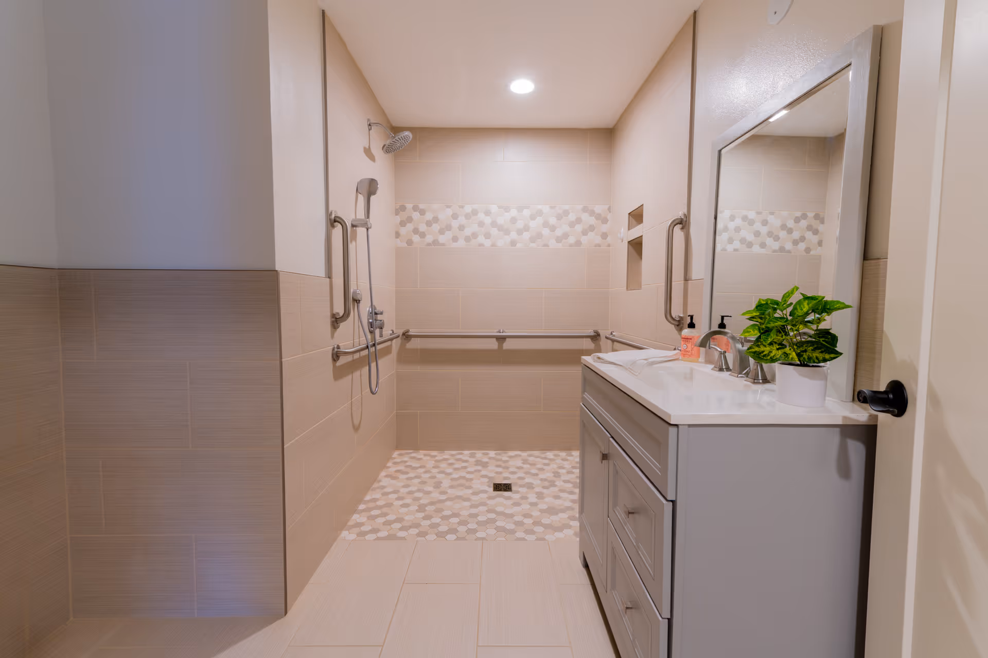 A modern bathroom with beige tiled walls and floor. The shower area features a handheld showerhead, a wall-mounted showerhead, and multiple grab bars for accessibility. The shower floor has a patterned tile design. To the right, there is a gray vanity with a white countertop, a sink, a mirror above it, a soap dispenser, and a small potted plant. The bathroom is well-lit with a ceiling light.