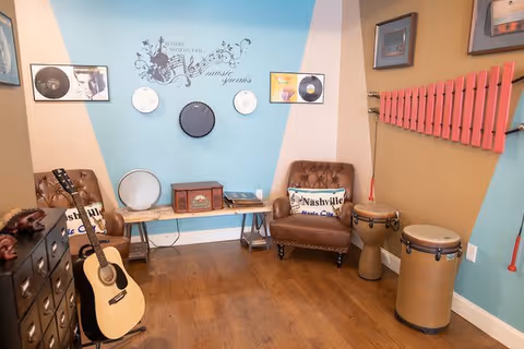 A cozy music-themed room with wooden flooring, two brown leather armchairs with Nashville Music City pillows, a wooden bench with various percussion instruments including tambourines and a drum, a guitar leaning against a black cabinet with drawers, a red xylophone mounted on the wall, and framed pictures and musical notes decor on the walls.